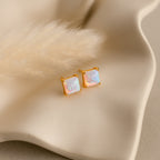 Princess Cut Opal Studs, crafted with a gold square design, rest on a wavy cream surface next to a soft beige plume, showcasing the vintage-inspired charm of these elegant earrings.