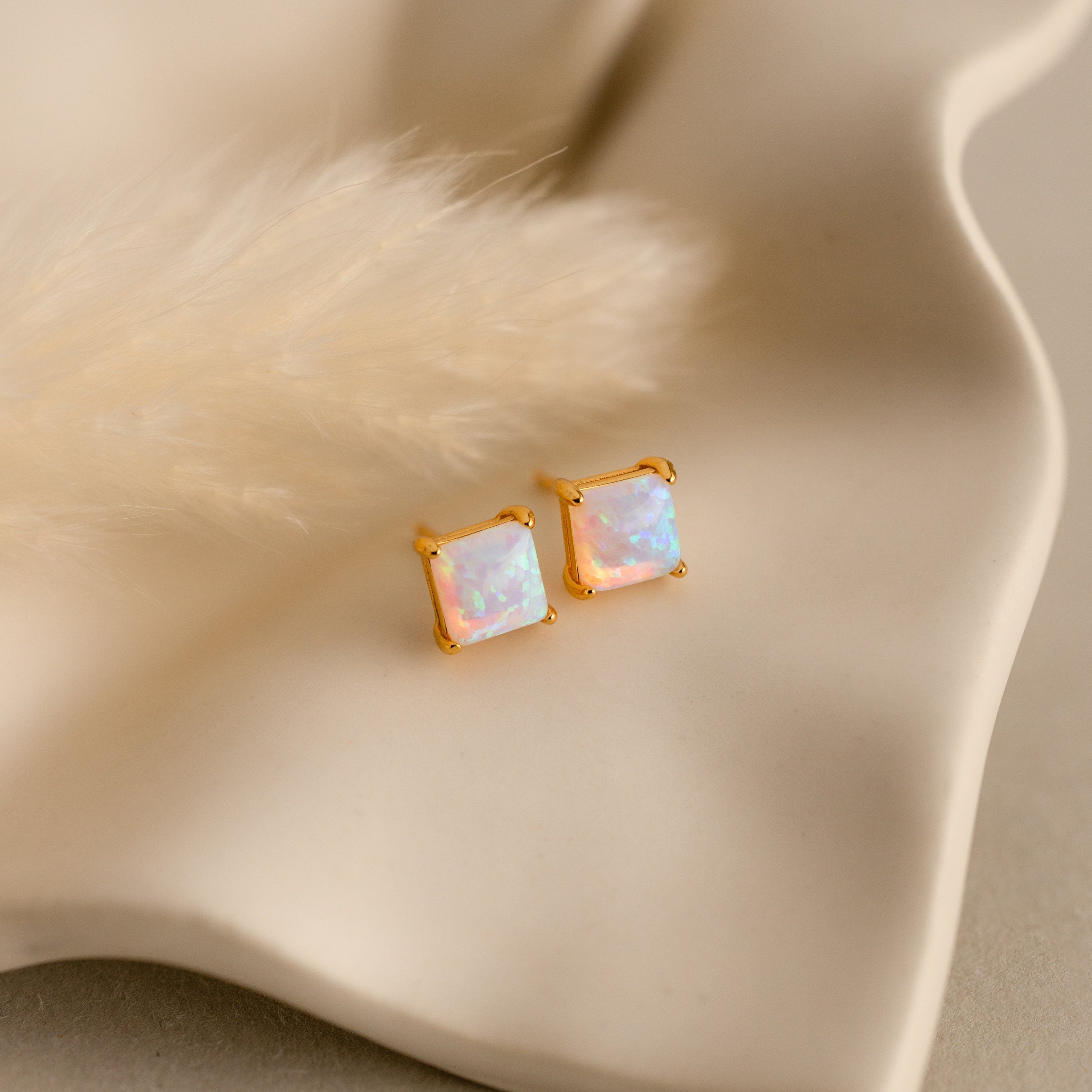 Princess Cut Opal Studs, crafted with a gold square design, rest on a wavy cream surface next to a soft beige plume, showcasing the vintage-inspired charm of these elegant earrings.