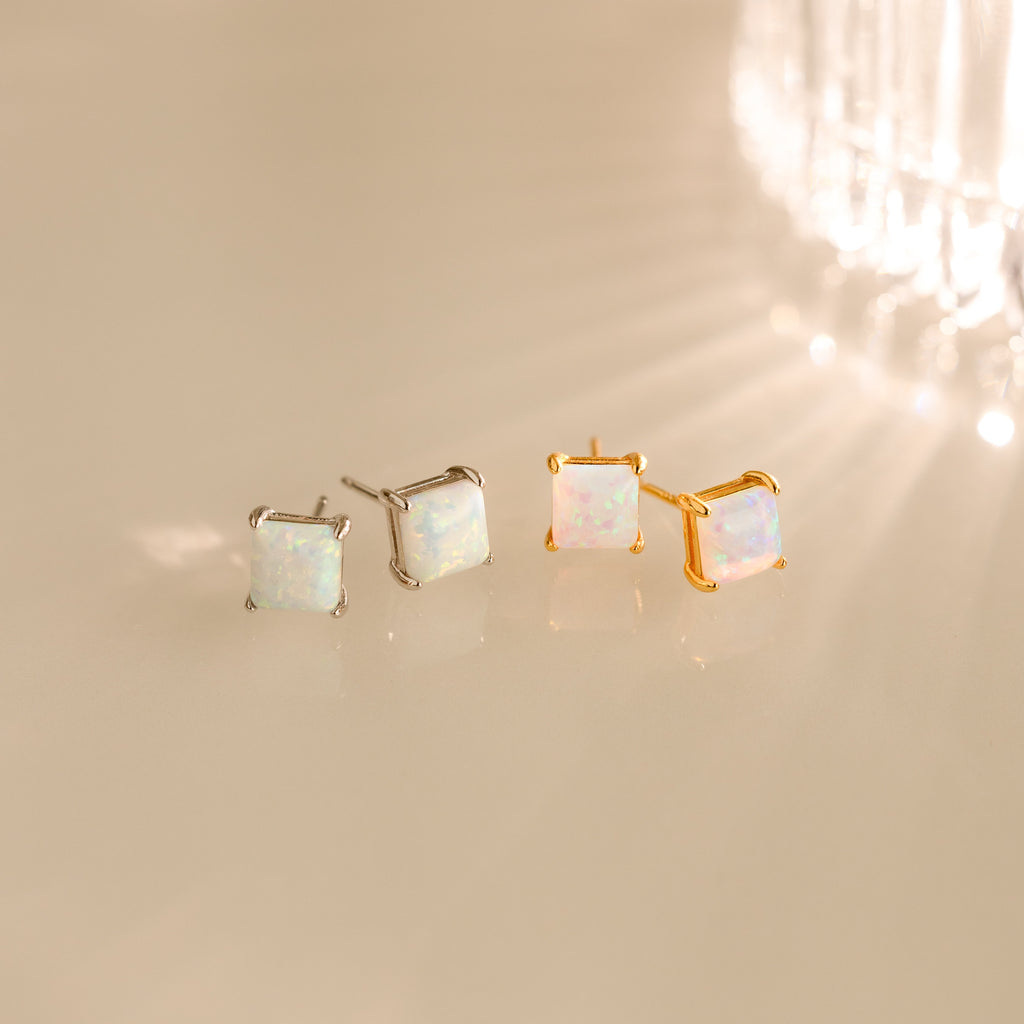 Two pairs of Princess Cut Opal Studs—one silver, one gold—bring vintage-inspired elegance as they shimmer on a reflective surface.