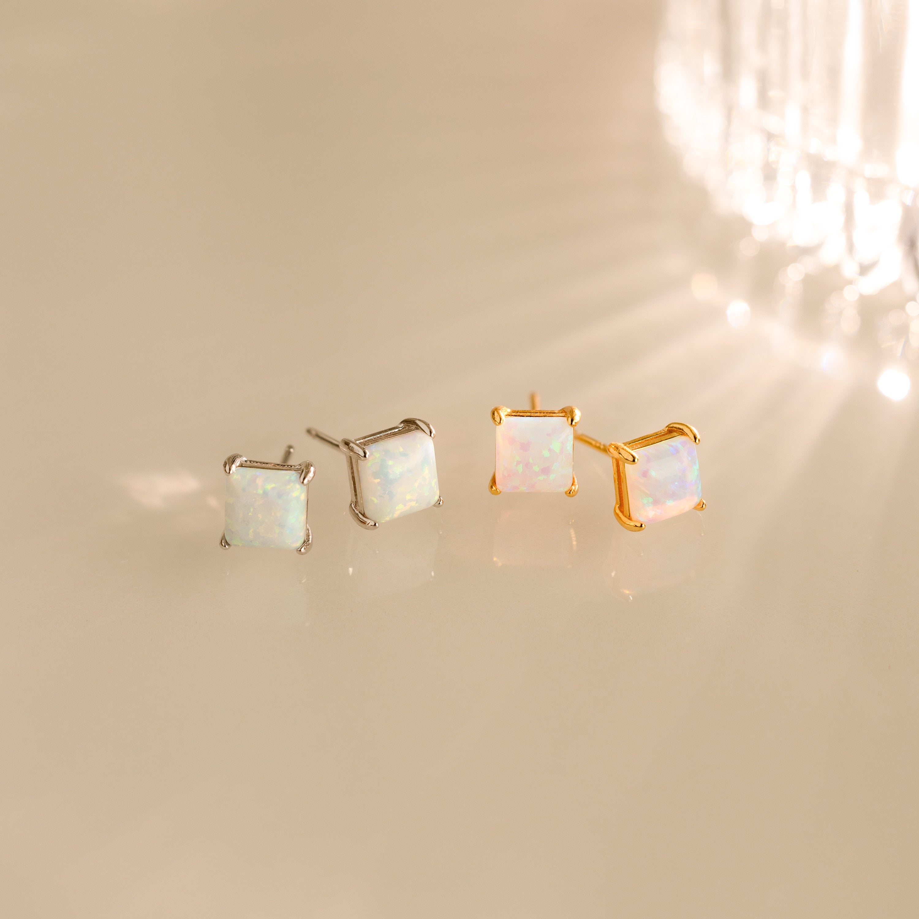 Two pairs of Princess Cut Opal Studs—one silver, one gold—bring vintage-inspired elegance as they shimmer on a reflective surface.