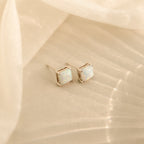 Princess Cut Opal Studs feature a timeless, vintage-inspired design with elegant silver settings, displayed on a soft beige fabric background.