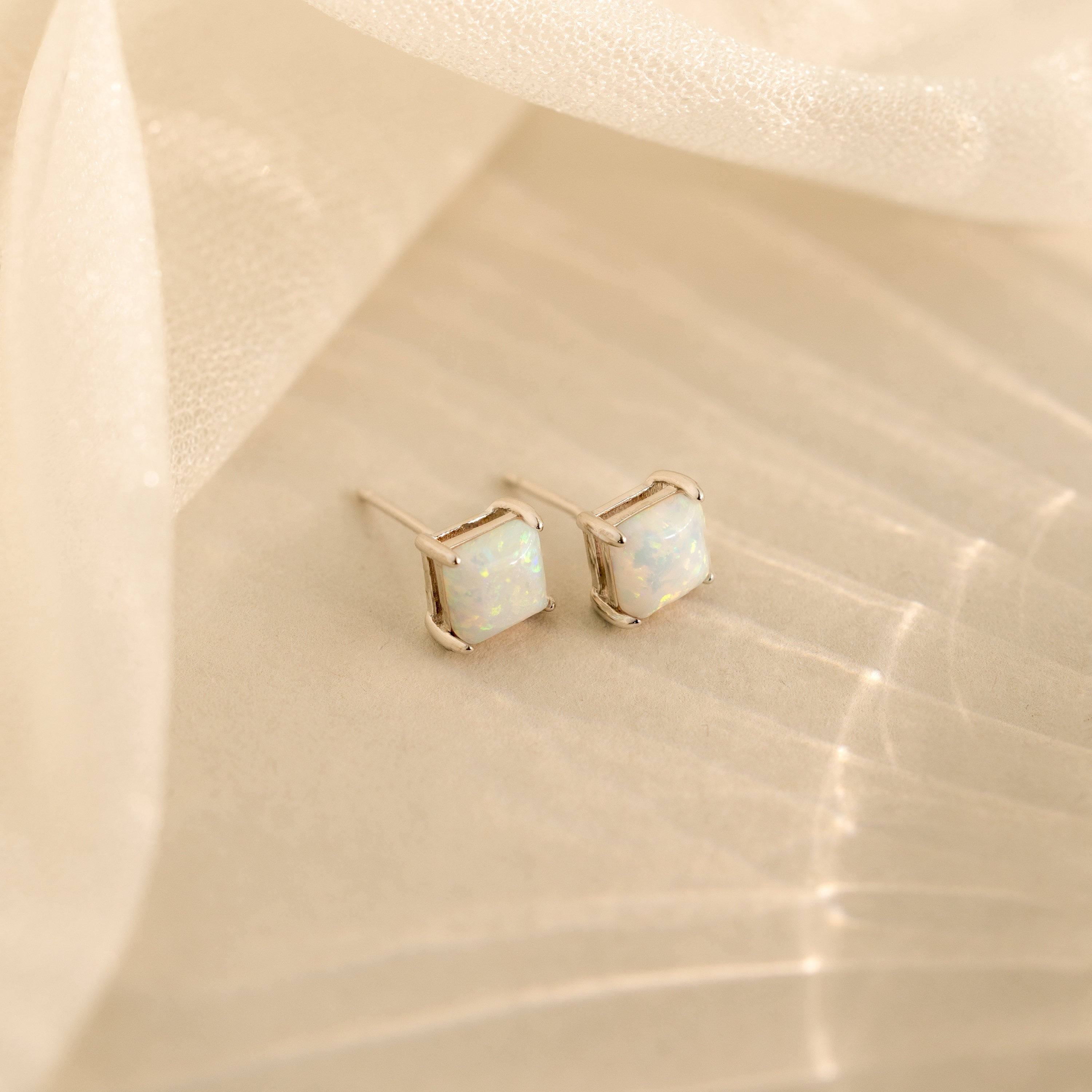 Princess Cut Opal Studs feature a timeless, vintage-inspired design with elegant silver settings, displayed on a soft beige fabric background.
