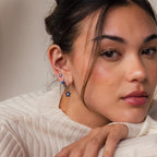 A woman with natural makeup, dressed in a cream blouse, rests her chin on her hand while wearing the Sapphire Flower Earrings Set in silver.