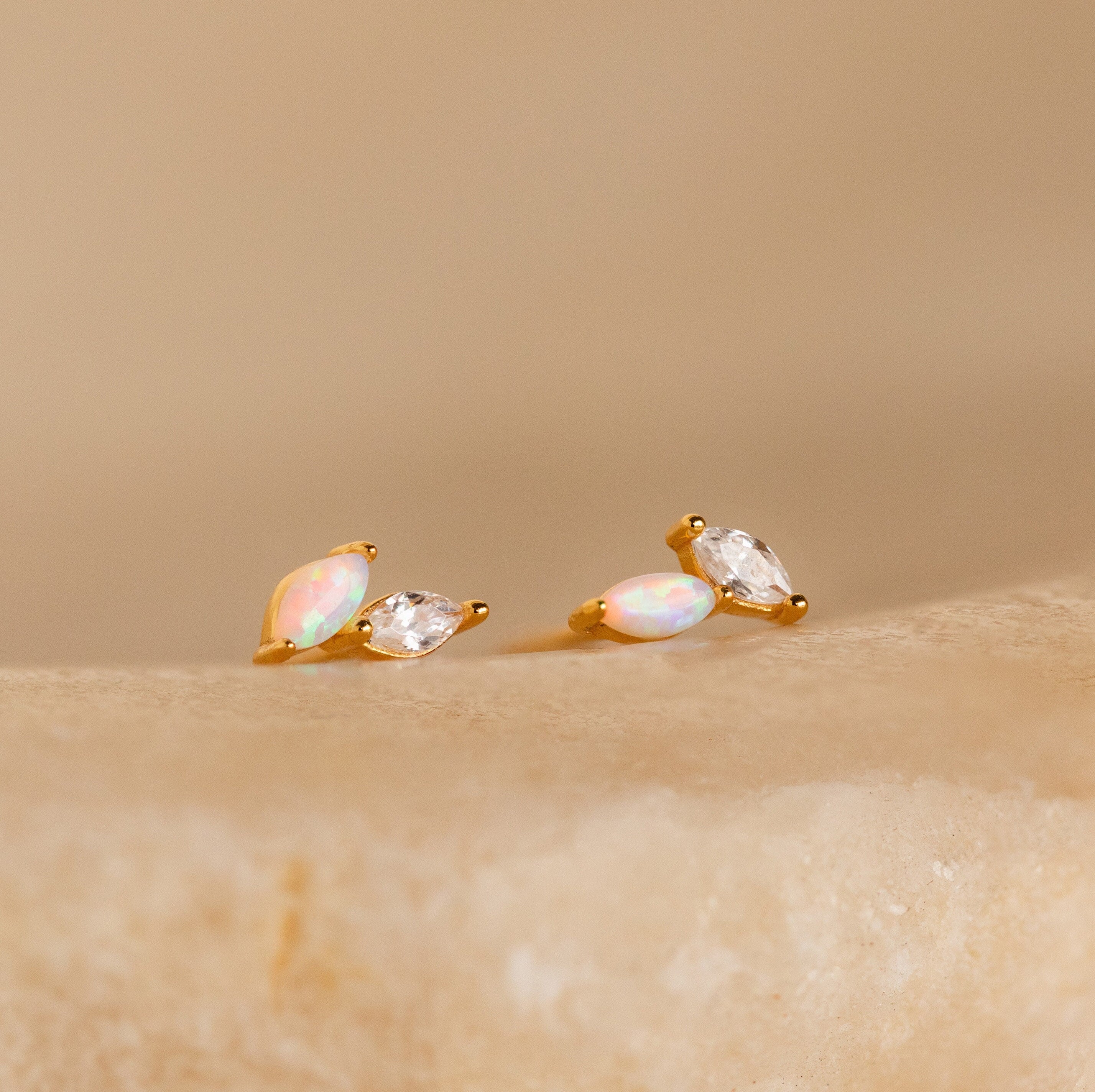 Two Opal & Diamond Marquise Studs, featuring shimmering opals and clear stones, rest on a beige stone surface—an ideal gift for bridesmaids or lovers of unique, mismatched earrings.