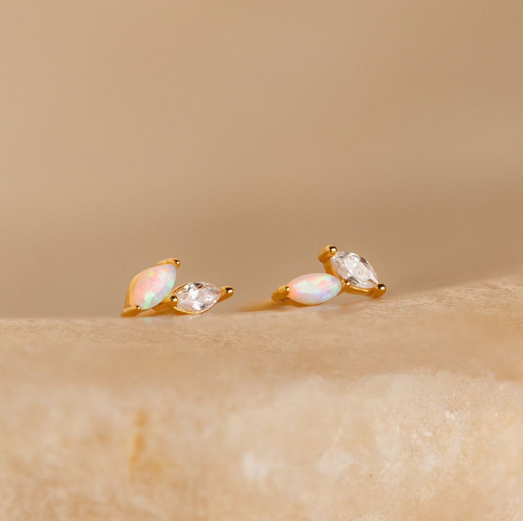 The Dainty Opal Stud Earrings Set features two small oval opals accented with marquise-cut clear stones on a beige, textured surface—ideal for minimalist jewelry lovers.