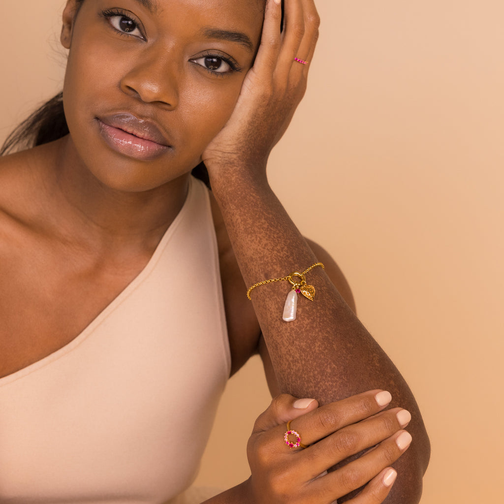 A woman in a beige top rests her face on her hand, wearing gold jewelry featuring pink stone accents and the Long Baroque Pearl Charm.