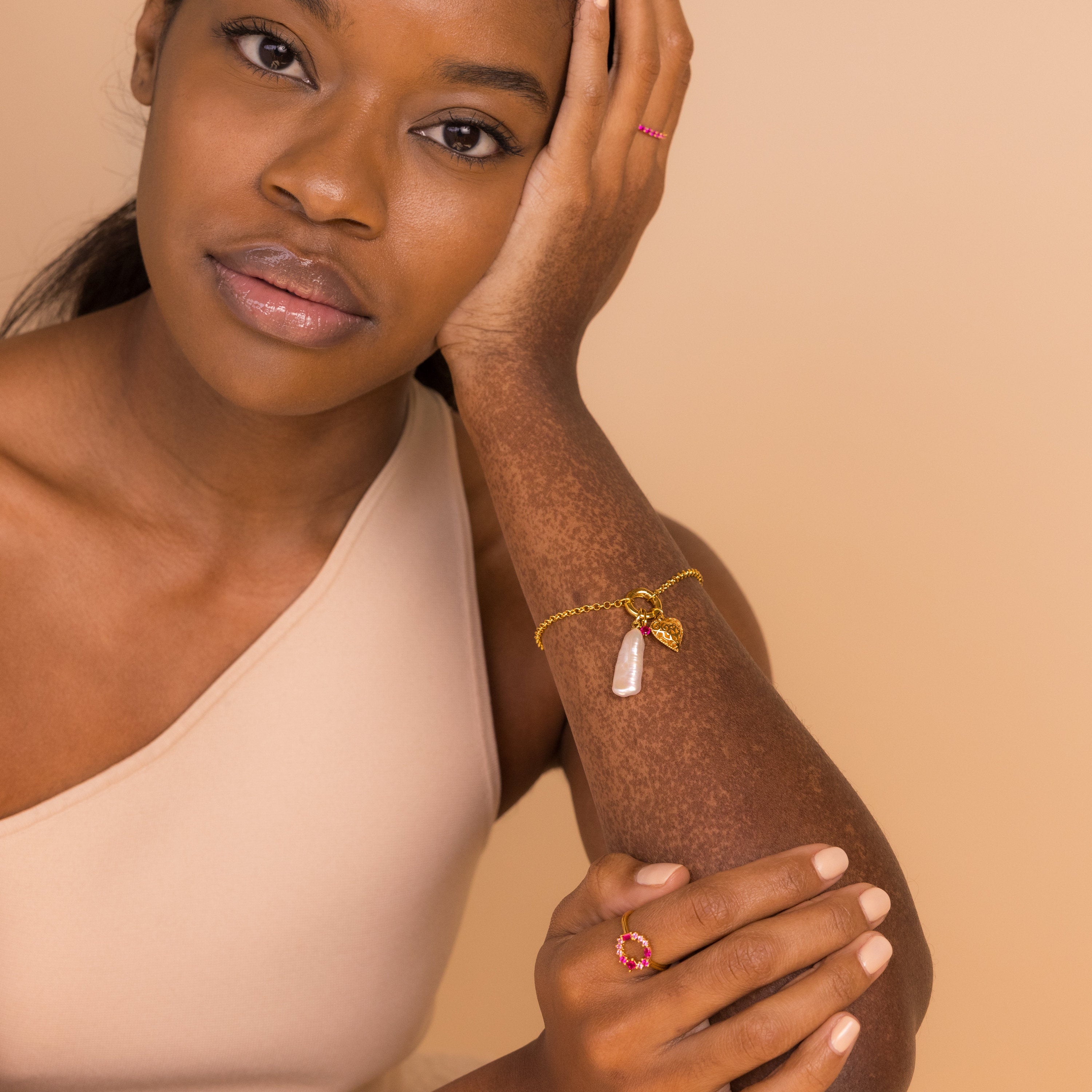 A woman in a beige top rests her face on her hand, wearing gold jewelry featuring pink stone accents and the Long Baroque Pearl Charm.