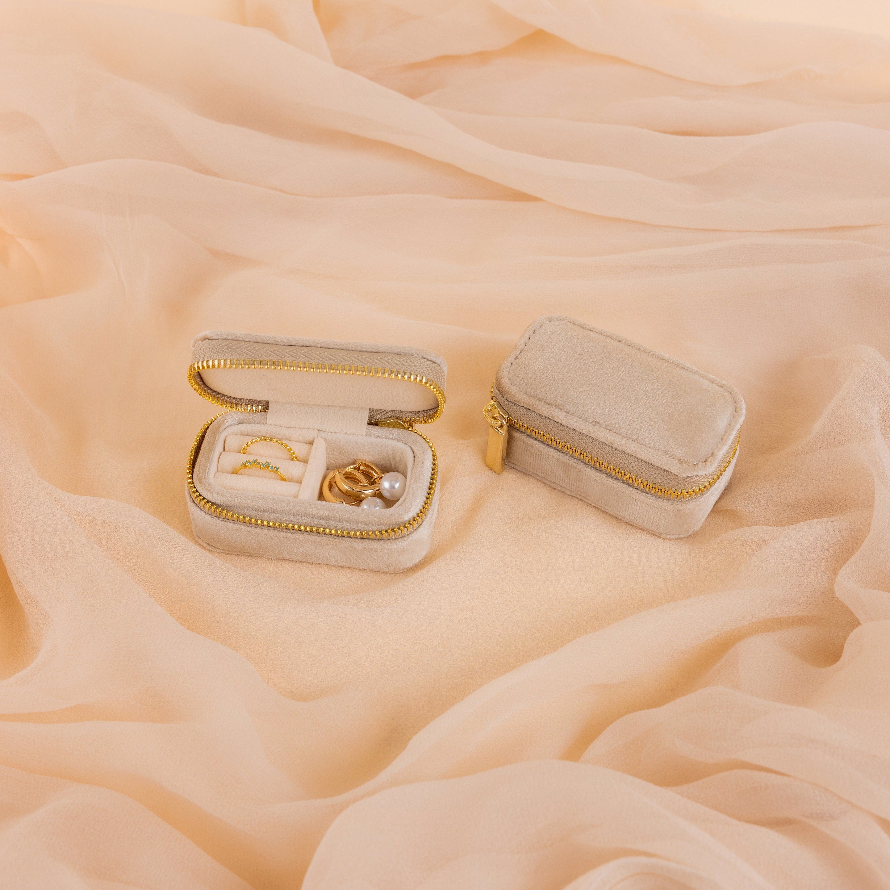 Two small beige Mini Travel Jewelry Boxes rest on peach fabric; one box is open, displaying rings and earrings—perfect for keeping your treasures organized wherever you go.