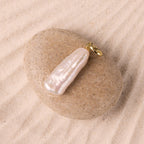 The Long Baroque Pearl Charm pendant is gracefully displayed on a smooth stone set atop soft sand.