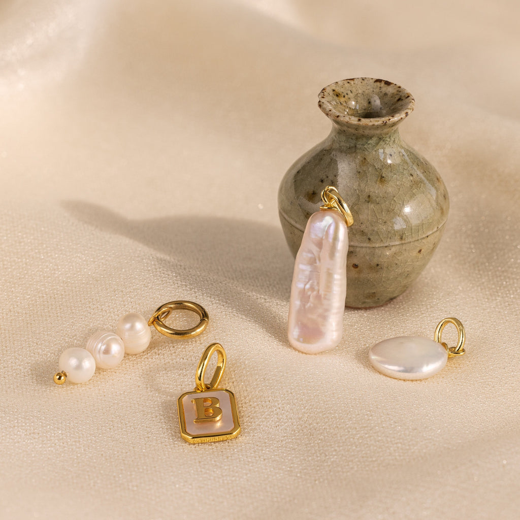 The Long Baroque Pearl Charm is showcased with gold and pearl pendants, arranged near a small ceramic vase on a beige fabric background.