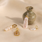 The Long Baroque Pearl Charm is showcased with gold and pearl pendants, arranged near a small ceramic vase on a beige fabric background.