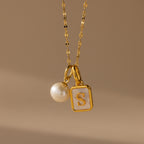 The Classic Pearl Charm in Sterling Silver is featured with a rectangular pendant engraved with the letter S on a beige background, paired elegantly with a delicate hoop earring for a refined look.