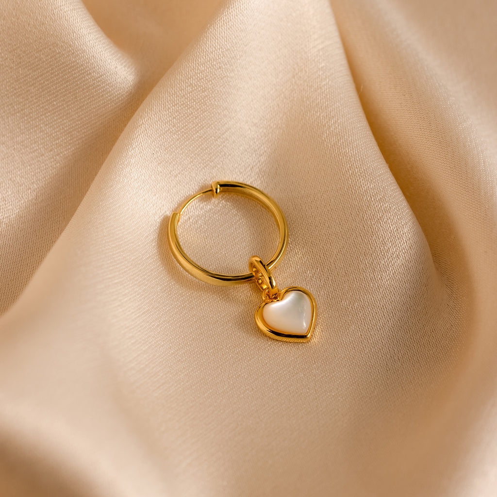 The Mother of Pearl Heart Charm, a gold hoop earring adorned with a delicate heart charm, rests on smooth beige satin—an elegant touch of vintage jewelry.