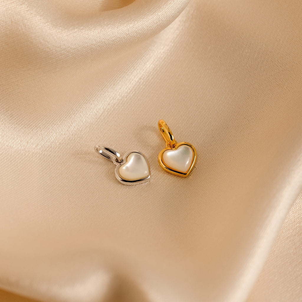 The Mother of Pearl Heart Charm—a timeless vintage piece—rests on smooth beige satin, its heart shape capturing elegant charm in silver and gold tones.