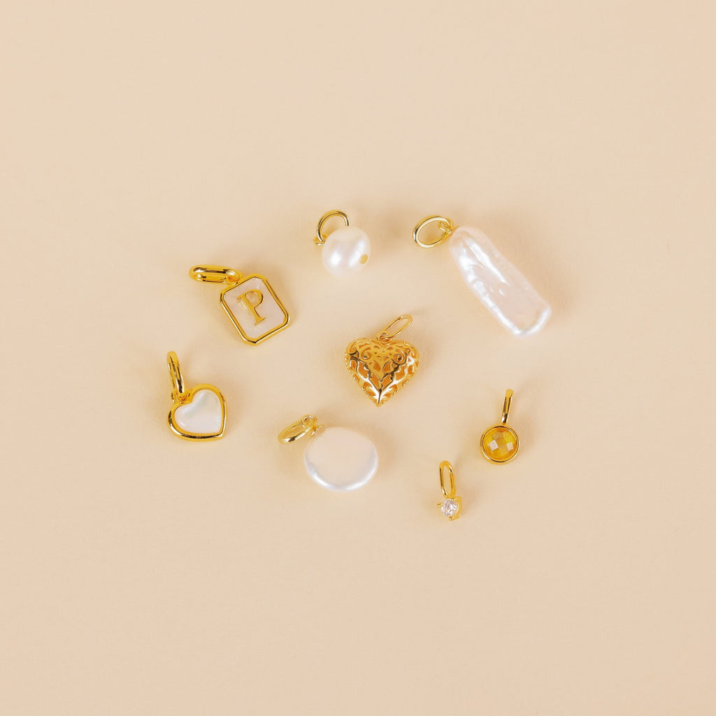 The Vintage Heart Charm in 18K Gold is featured with gold and pearl charms, a rectangle with P, and round pendants on a beige background—a lovely addition to any jewelry collection.