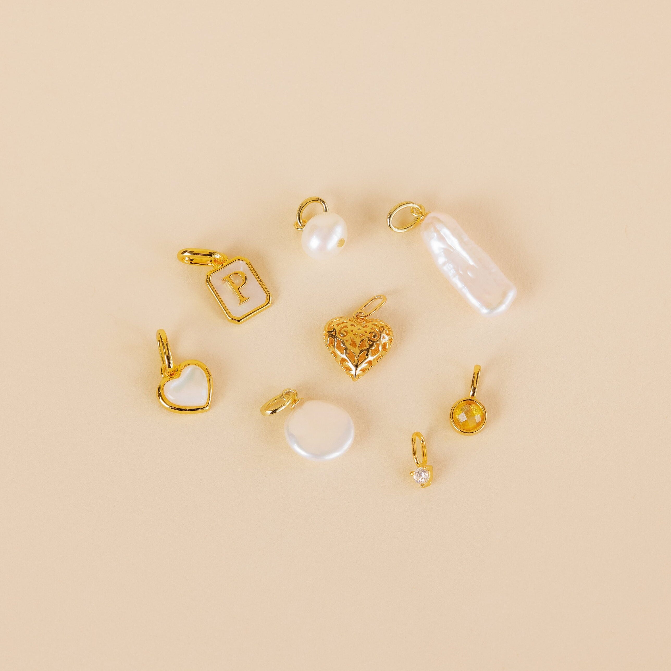 The Vintage Heart Charm in 18K Gold is featured with gold and pearl charms, a rectangle with P, and round pendants on a beige background—a lovely addition to any jewelry collection.