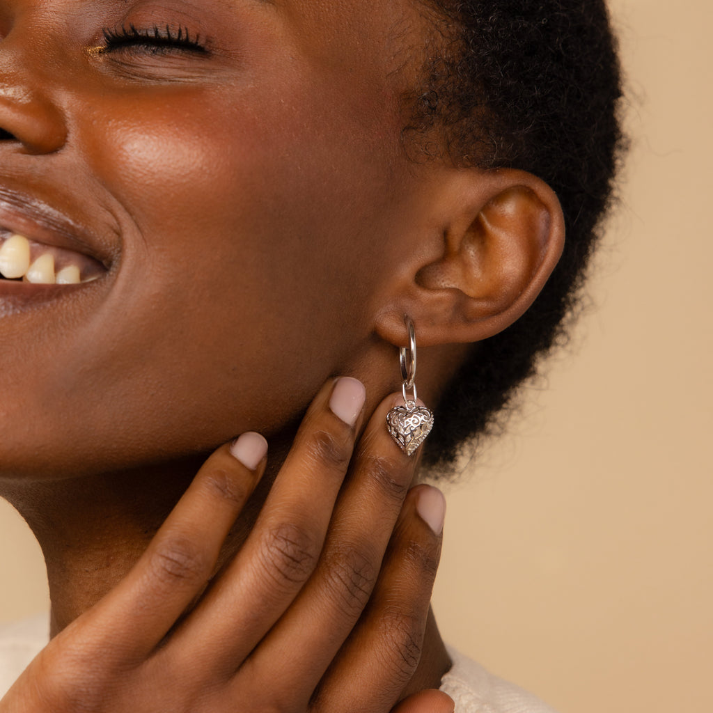 Smiling woman touches her face, wearing the Vintage Heart Charm in Sterling Silver earring against a beige background—a stunning addition to any jewelry collection.