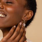 Smiling woman touches her face, wearing the Vintage Heart Charm in Sterling Silver earring against a beige background—a stunning addition to any jewelry collection.
