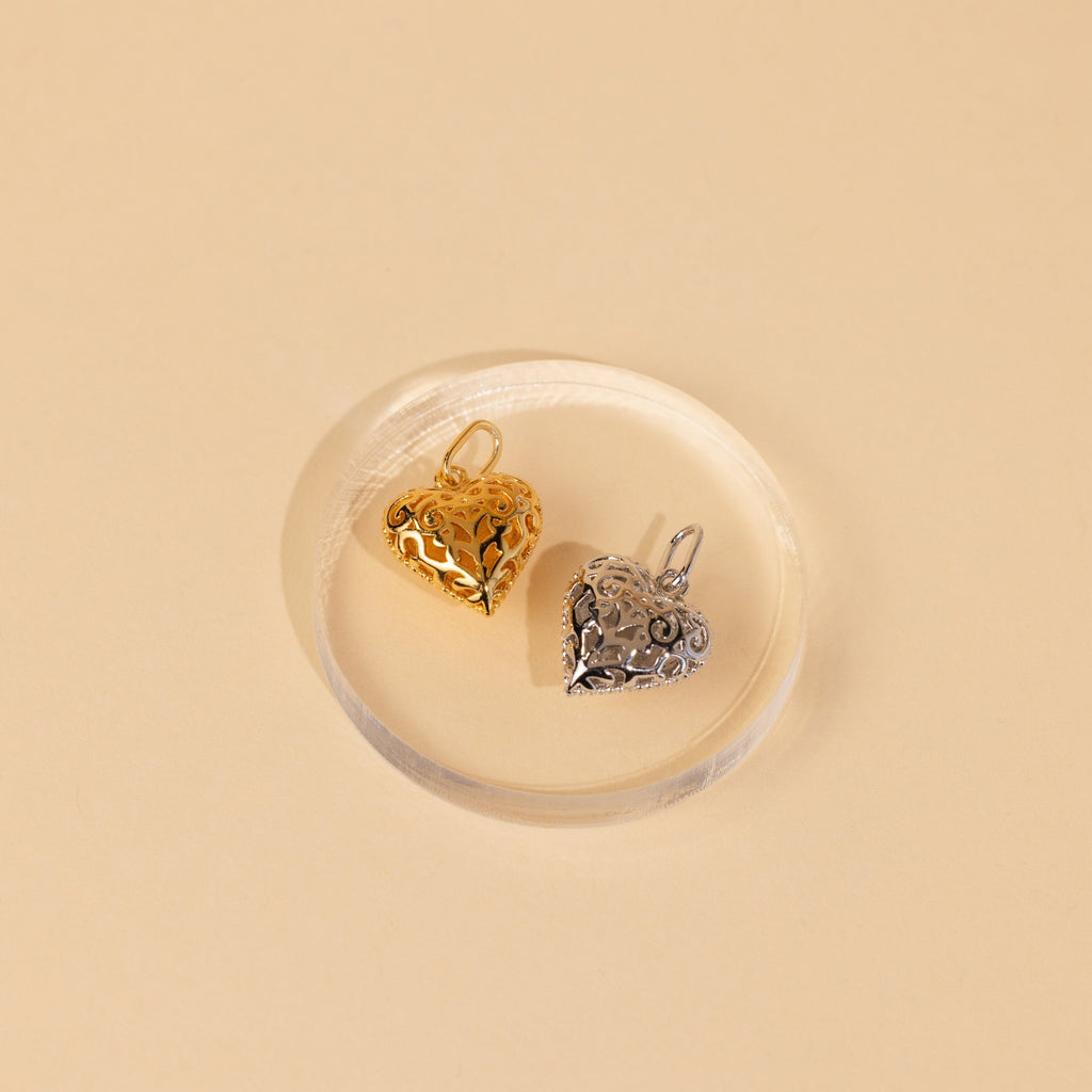 The Vintage Heart Charm in Sterling Silver features a filigree design and rests on a clear round dish against a beige background—perfect as an anniversary gift or a unique addition to your jewelry collection.