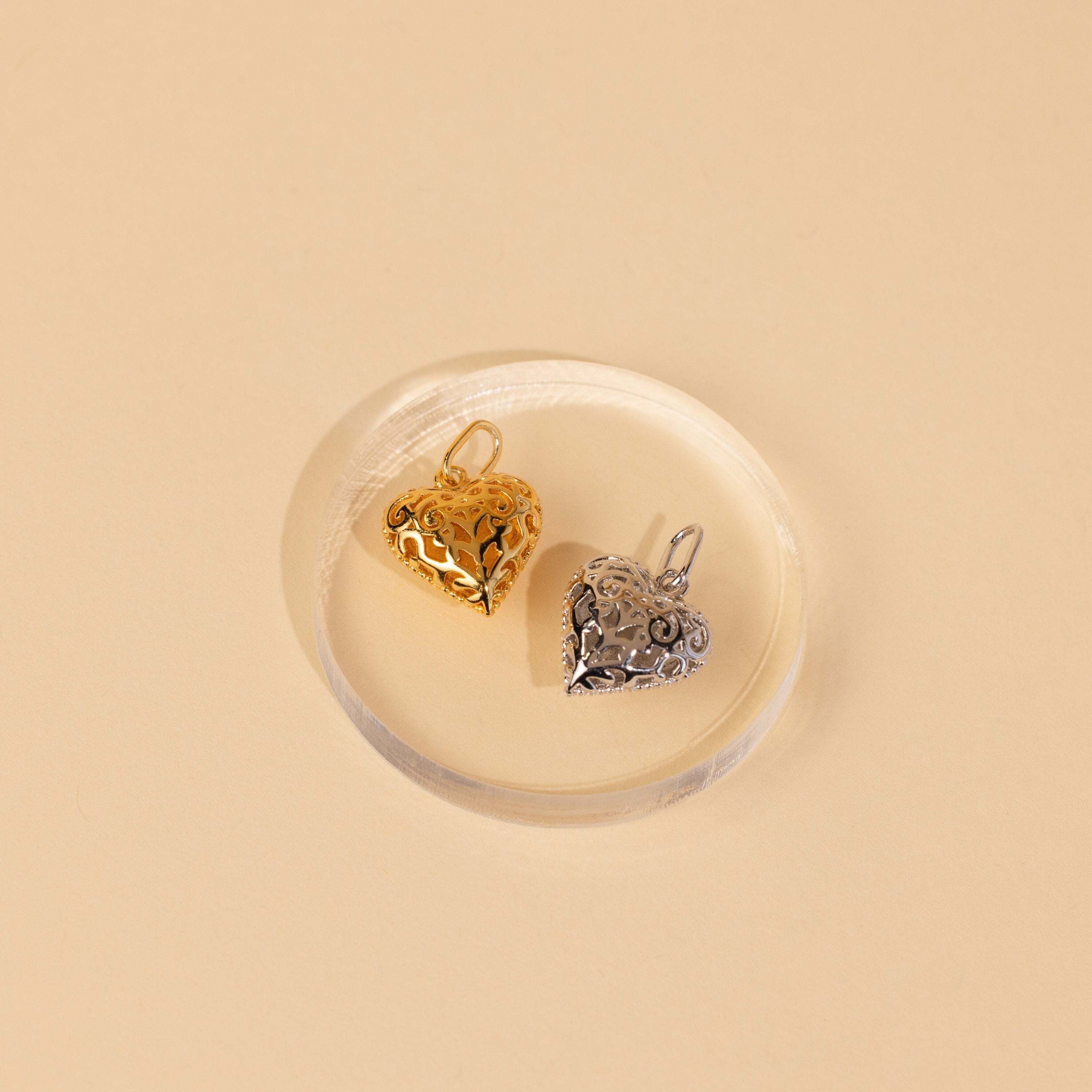 The Vintage Heart Charm in Sterling Silver features a filigree design and rests on a clear round dish against a beige background—perfect as an anniversary gift or a unique addition to your jewelry collection.