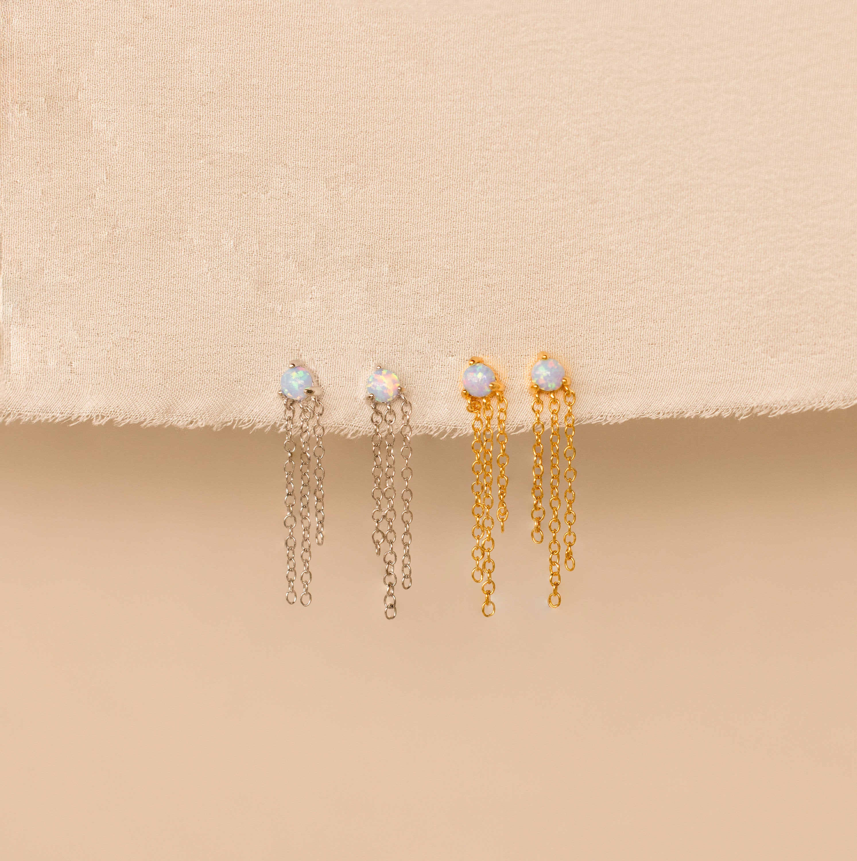 Opal Chain Earrings in both silver and gold are displayed on beige fabric, featuring stud opals with dangling chains—a beautiful addition to any jewelry collection.