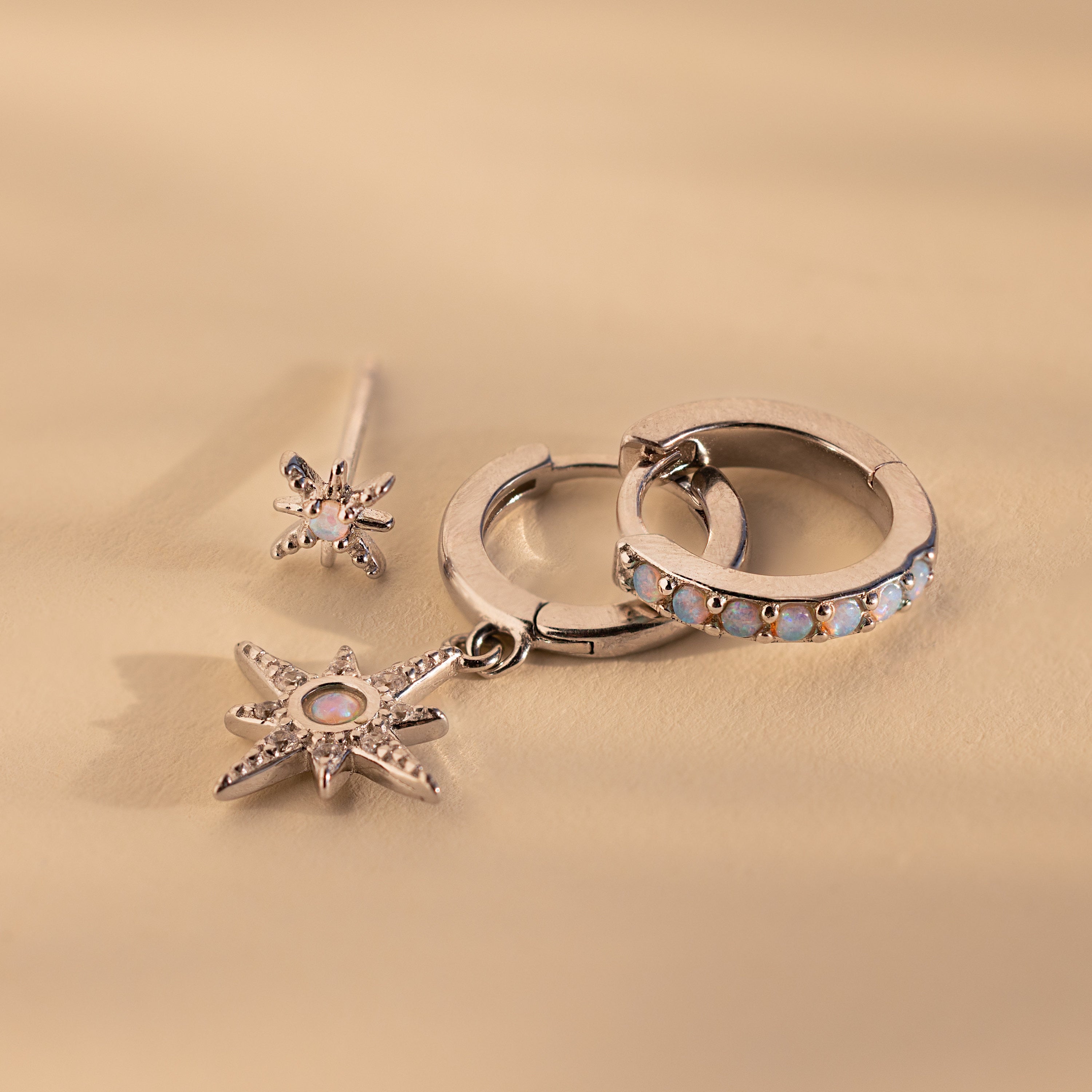 The Opal Starburst Earrings Set, featuring three silver earrings with star and gemstone details, is displayed on a beige background for a dazzling effect.