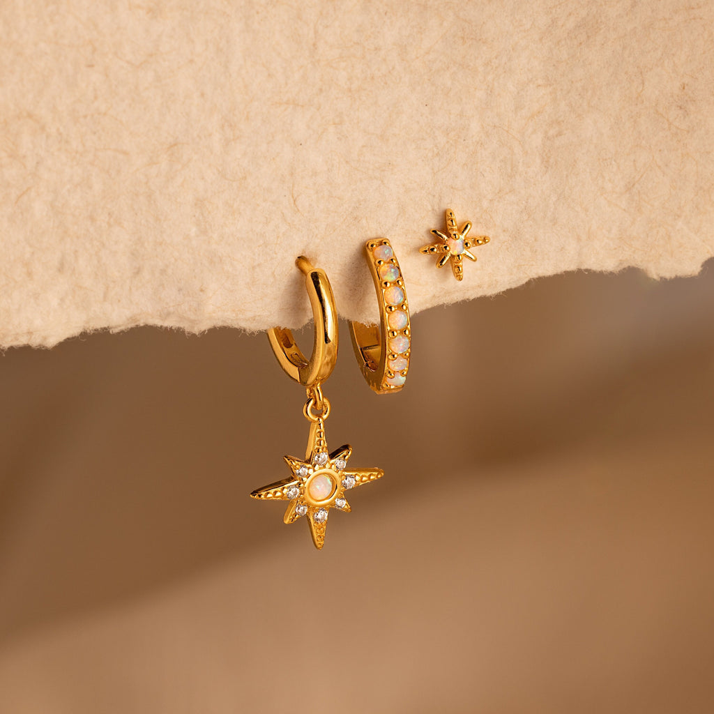 The Opal Starburst Earrings Set showcases three gold earrings with star and pearl-like details, elegantly displayed on textured beige paper—a stunning addition to your jewelry collection.