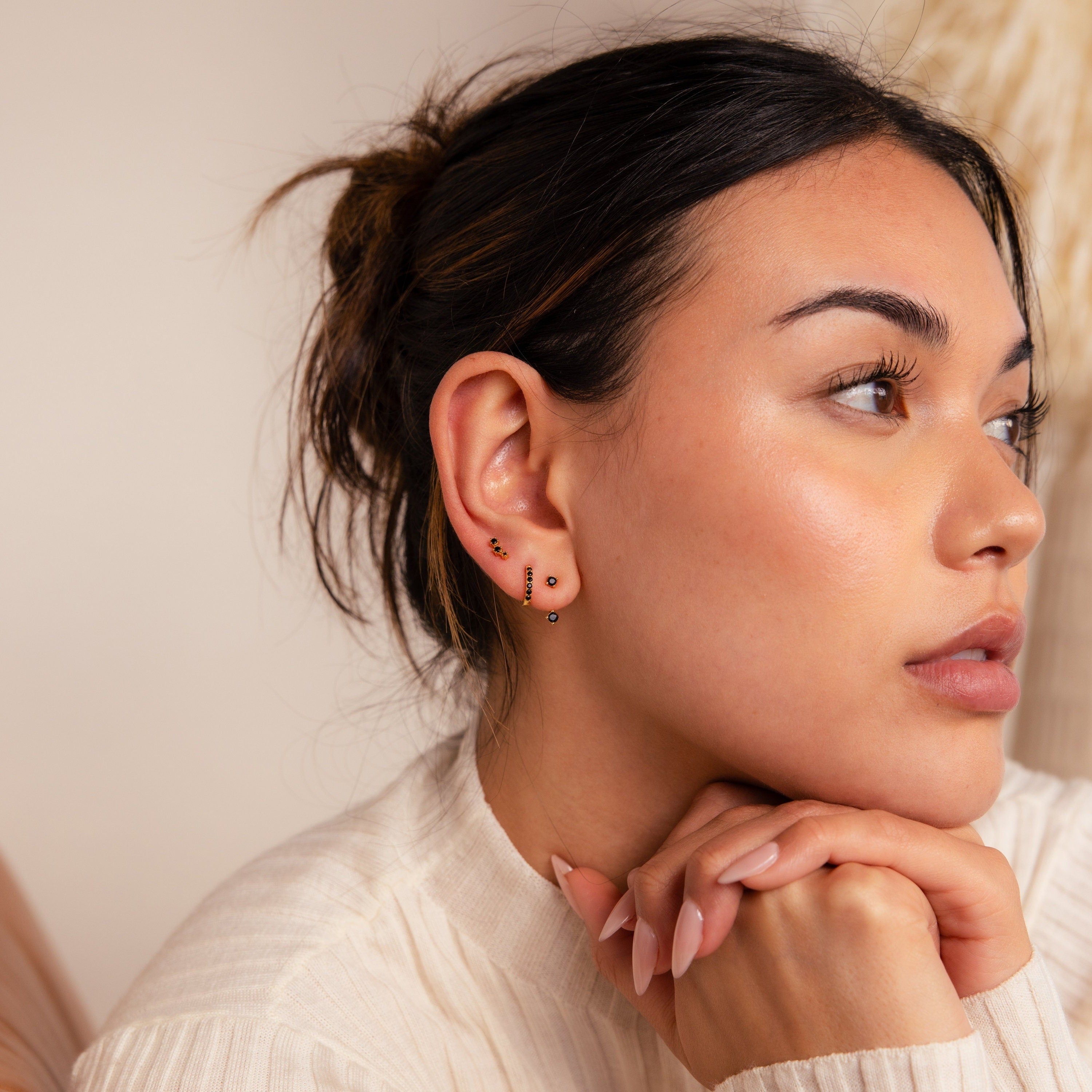 A woman with smooth skin wears the Black Diamond Earrings Set, including a climber stud, as she rests her chin on her hands and gazes to the right.