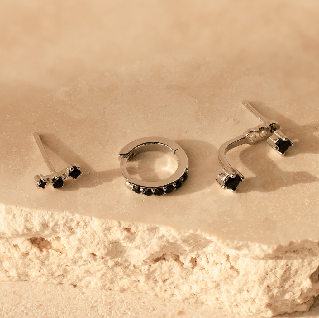 The Black Diamond Earrings Set features three pieces of minimalist silver jewelry with black stones, displayed on a textured beige stone surface.