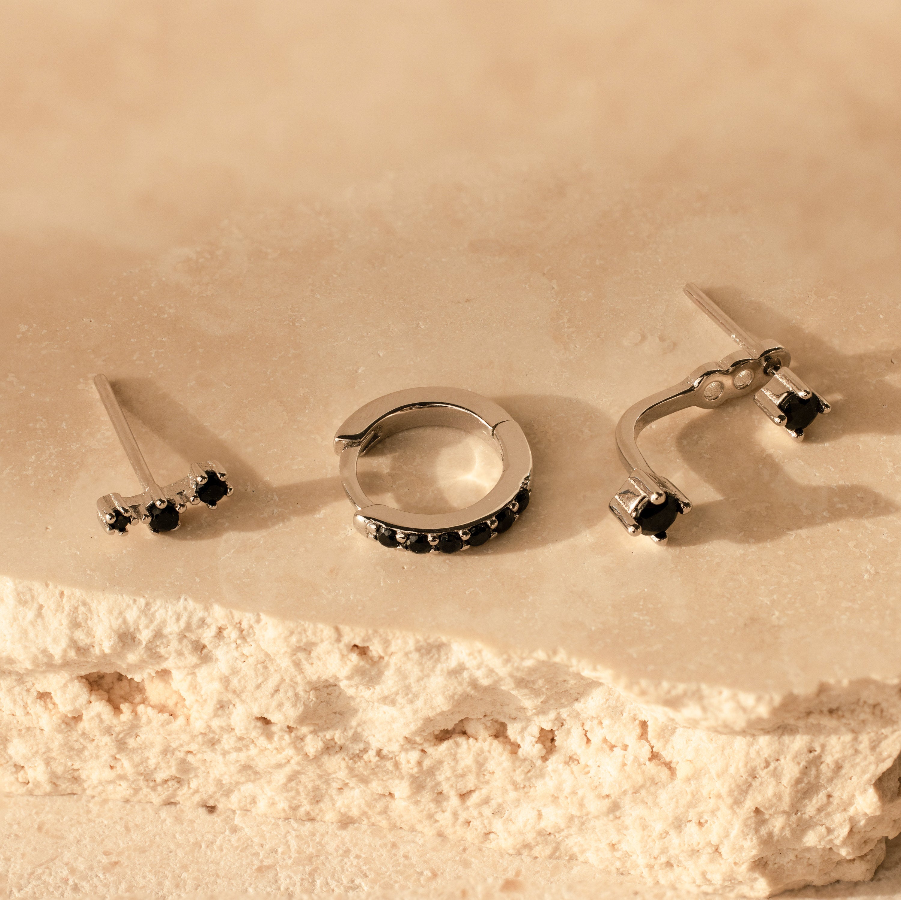 The Black Diamond Earrings Set features three pieces of minimalist silver jewelry with black stones, displayed on a textured beige stone surface.