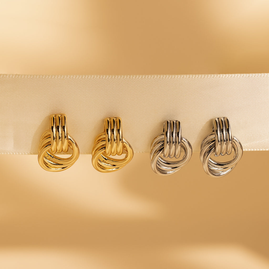 Four Triple Knot Earrings—two gold and two silver—displayed along a cream-colored ribbon.