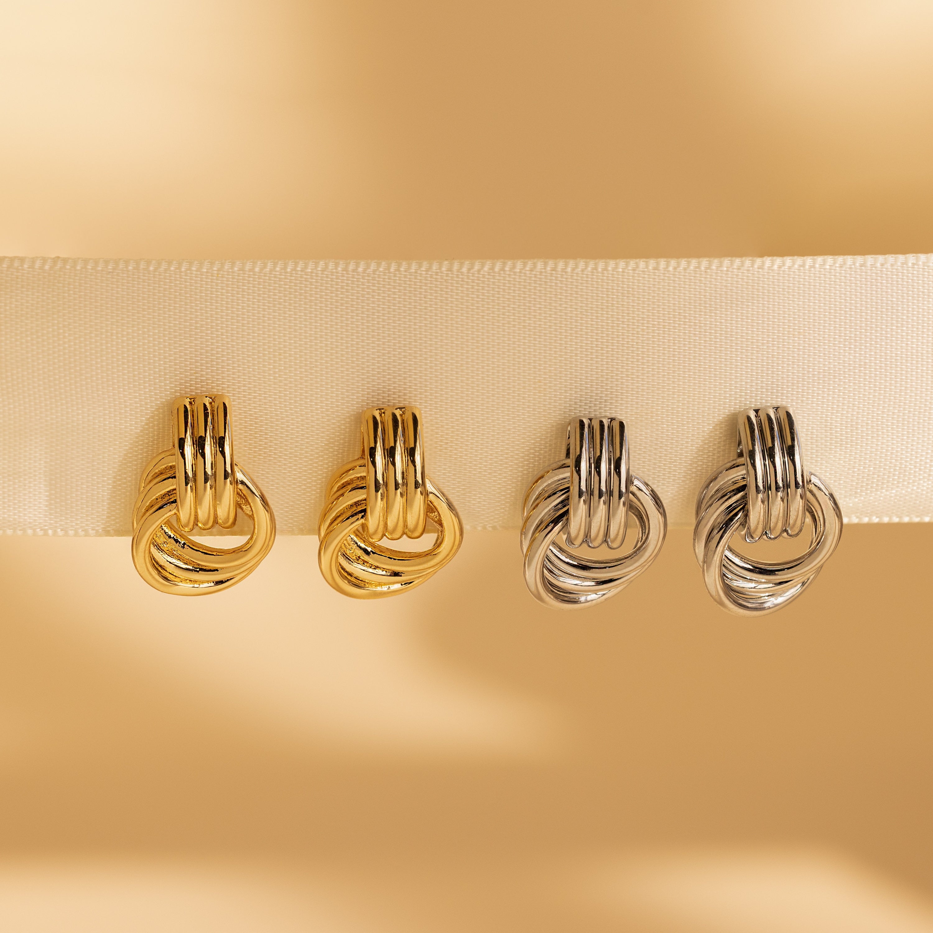 Four Triple Knot Earrings—two gold and two silver—displayed along a cream-colored ribbon.