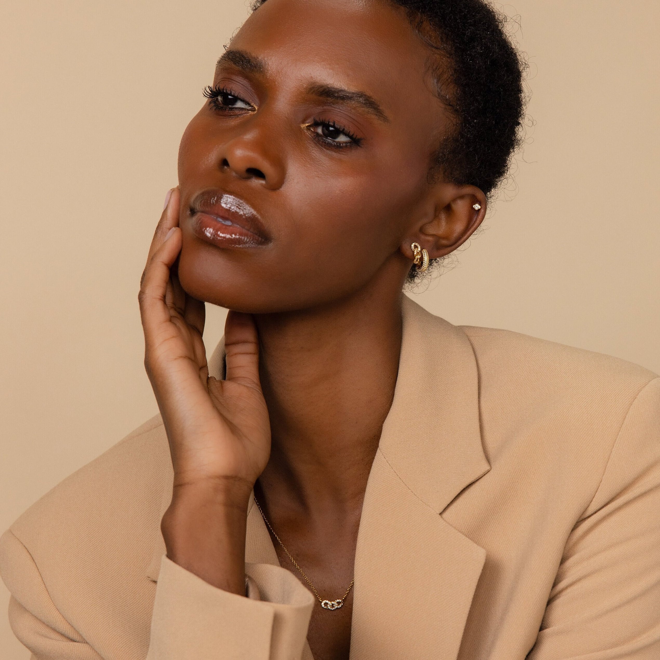 Woman in a beige blazer poses with her hand on her face, wearing elegant Pave Link Studs and a sparkling diamond necklace.