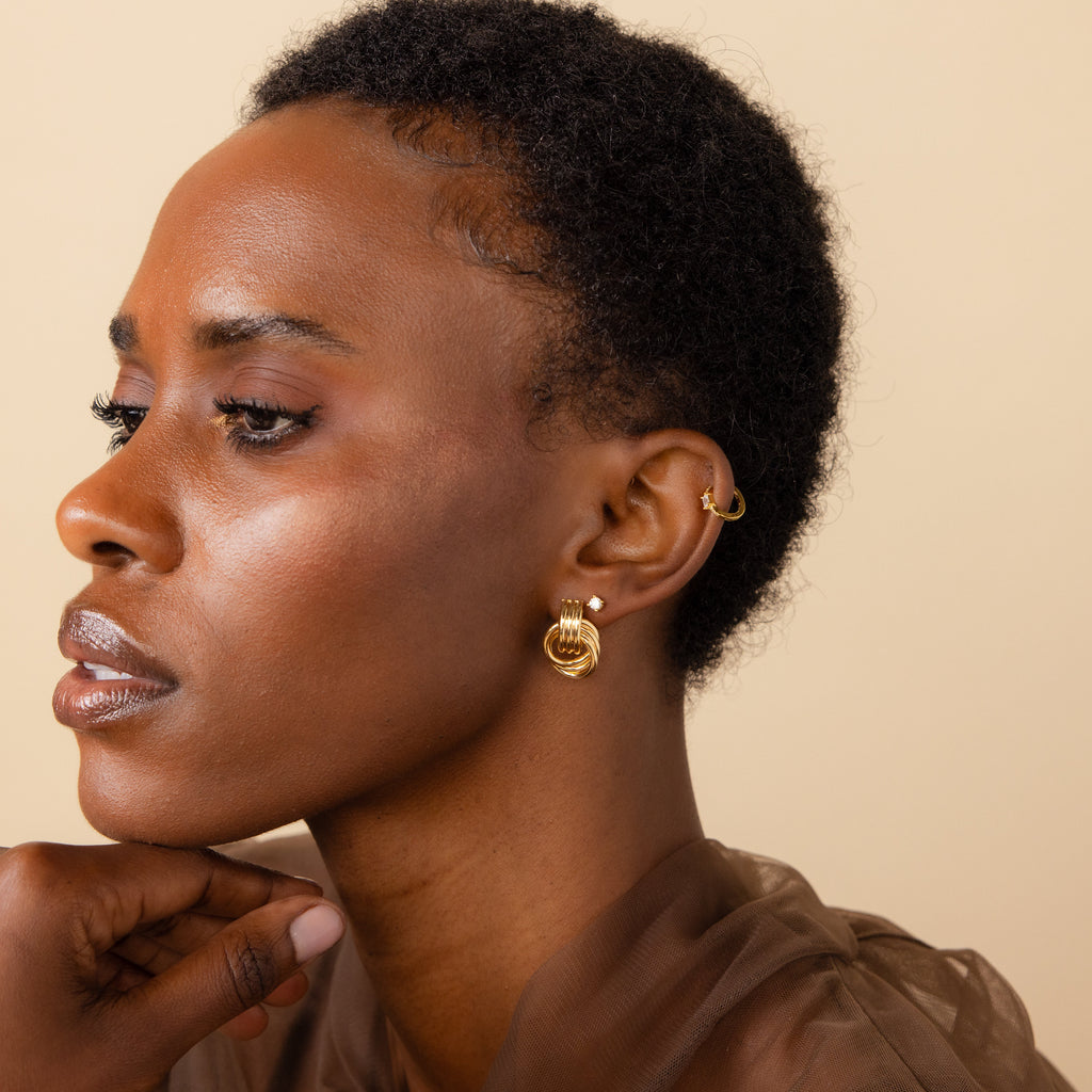 A woman with short hair wears the elegant Triple Knot Earrings and a brown sheer top, gazing thoughtfully to the side.