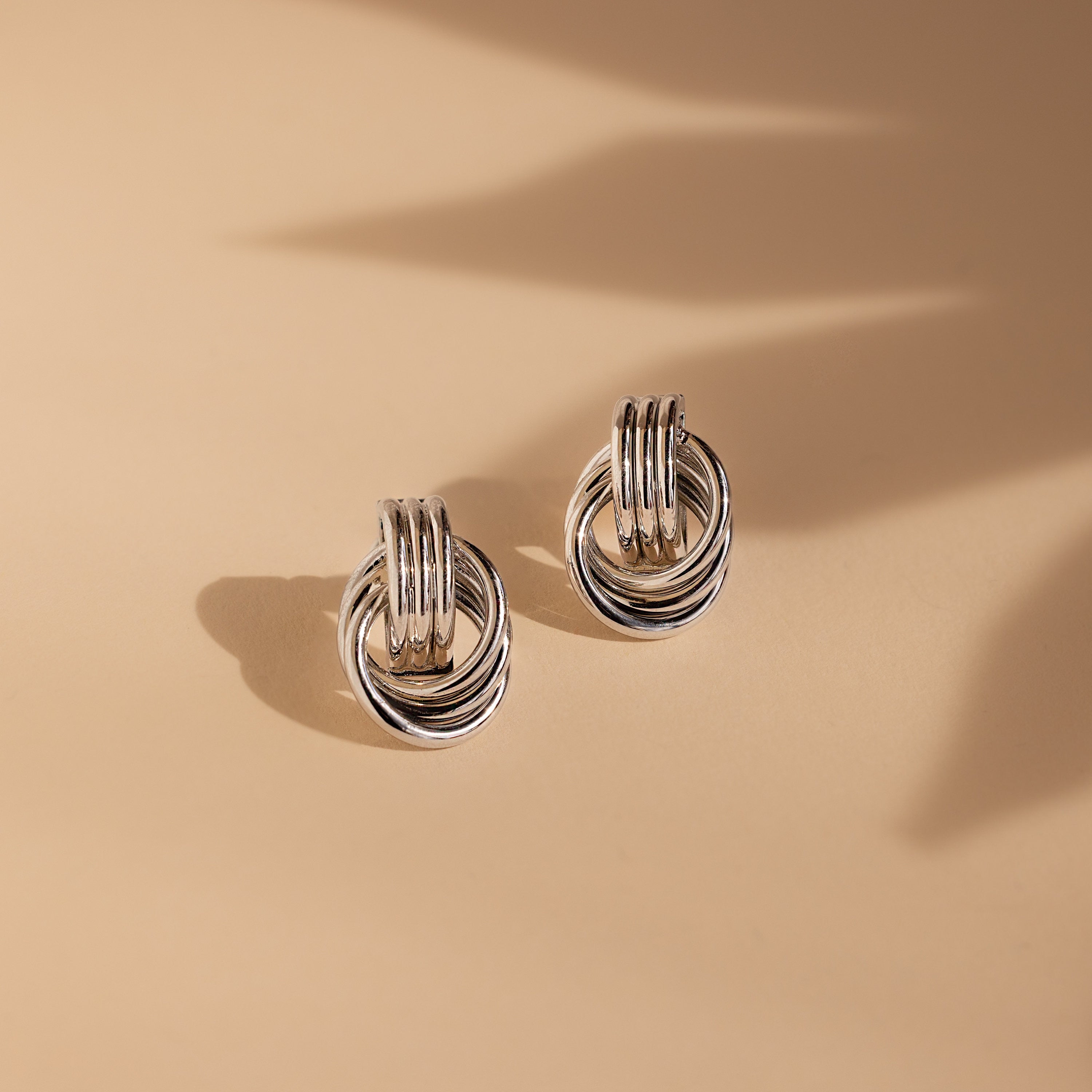 Triple Knot Earrings: Stylish silver hoop stud earrings, ideal for an anniversary gift, showcased on a beige background with soft shadows.