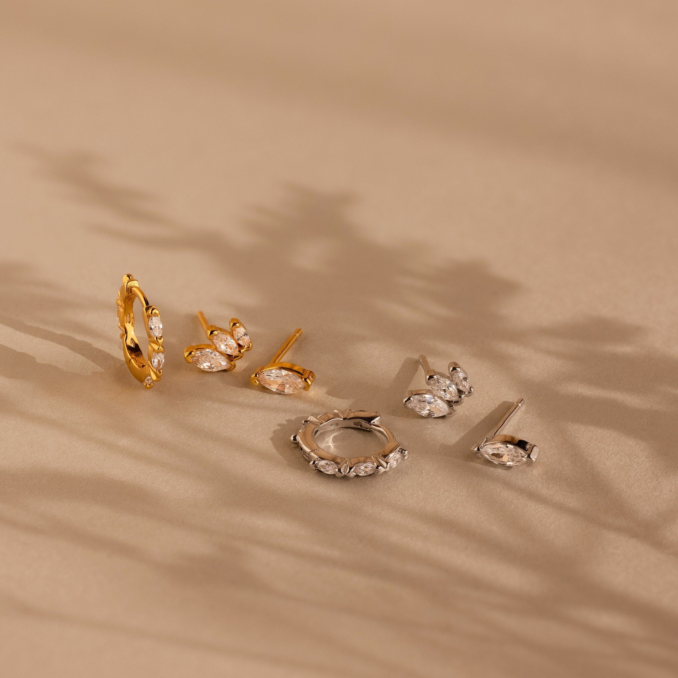 The Marquise Diamond Earrings Set, featuring gold and silver with gemstones, is displayed on a beige surface, exuding wedding elegance amid soft shadows in the background.