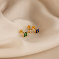 The Flower Flat Back Studs Set features three gold flower stud earrings with green, clear, and blue stones, displayed on soft beige fabric—ideal for comfortable flat back wear.