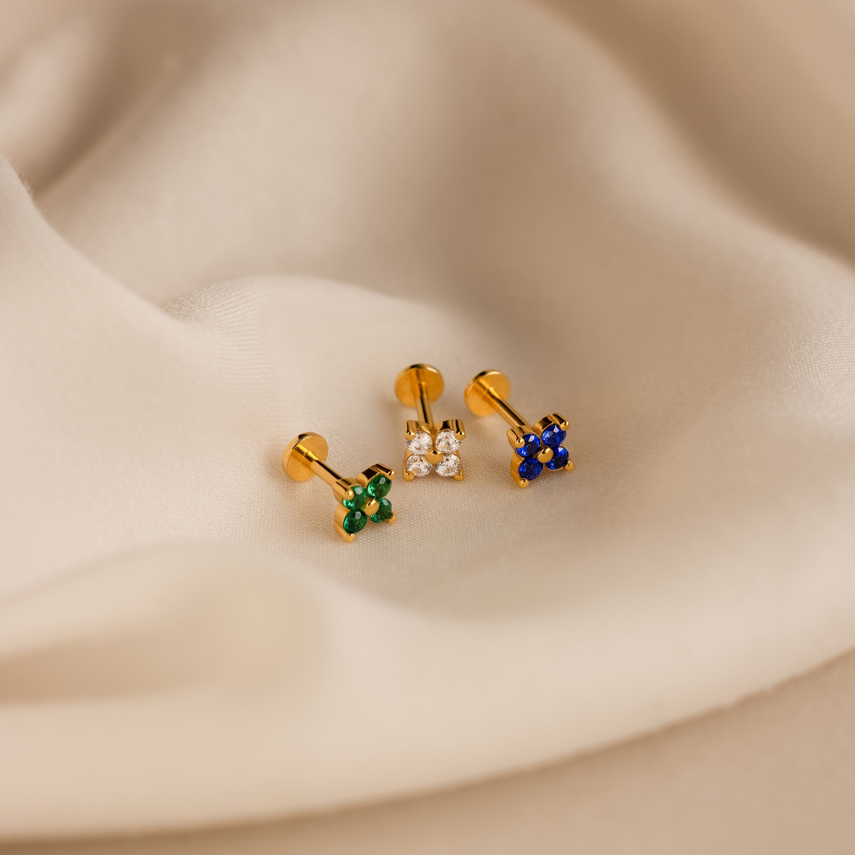 The Flower Flat Back Studs Set features three gold flower stud earrings with green, clear, and blue stones, displayed on soft beige fabric—ideal for comfortable flat back wear.