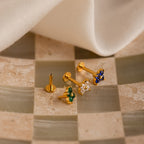 The Flower Flat Back Studs Set features three gold flower earrings with green, white, and blue stones, displayed on a tiled surface near white fabric. These elegant flat back studs enhance any look.