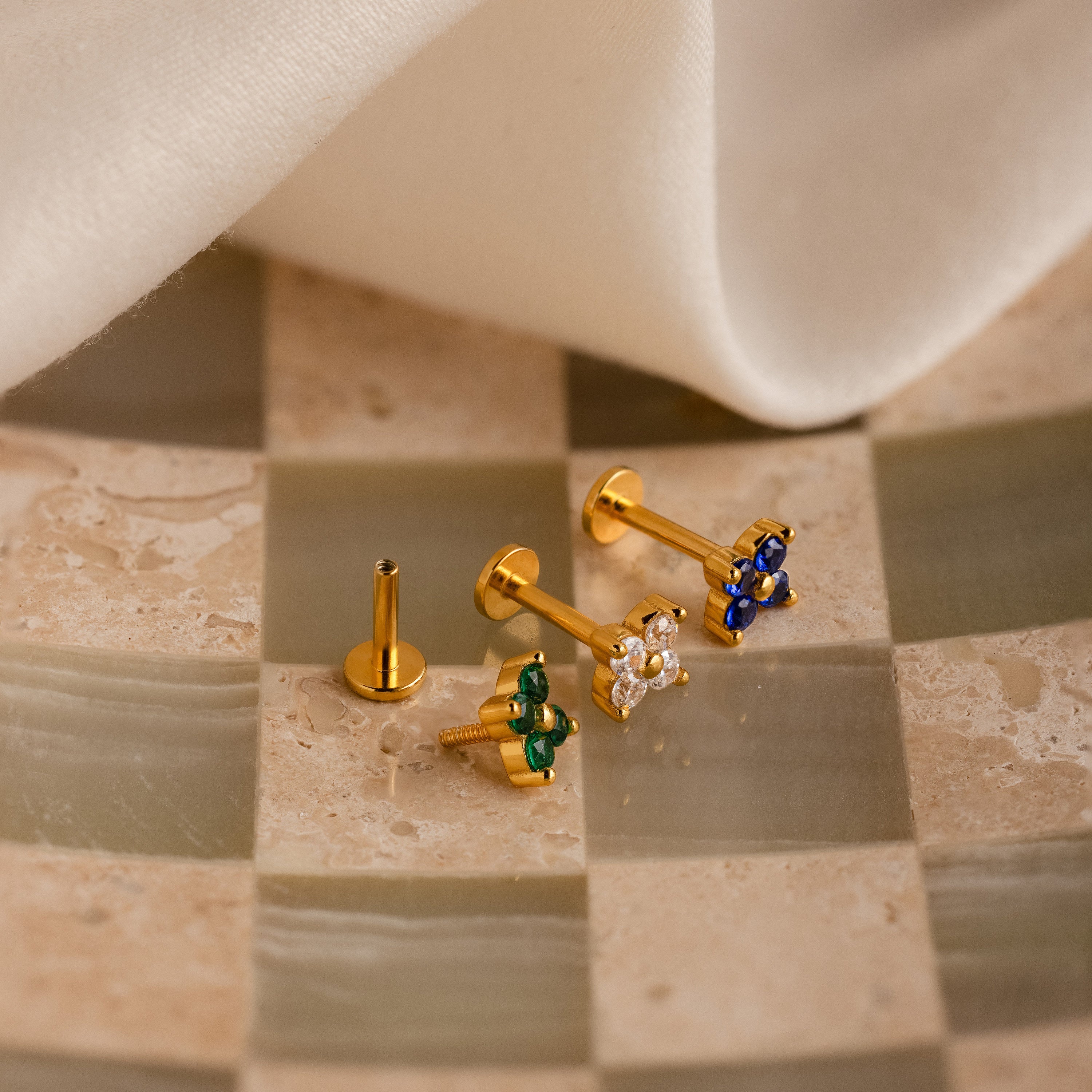 The Flower Flat Back Studs Set features three gold flower earrings with green, white, and blue stones, displayed on a tiled surface near white fabric. These elegant flat back studs enhance any look.