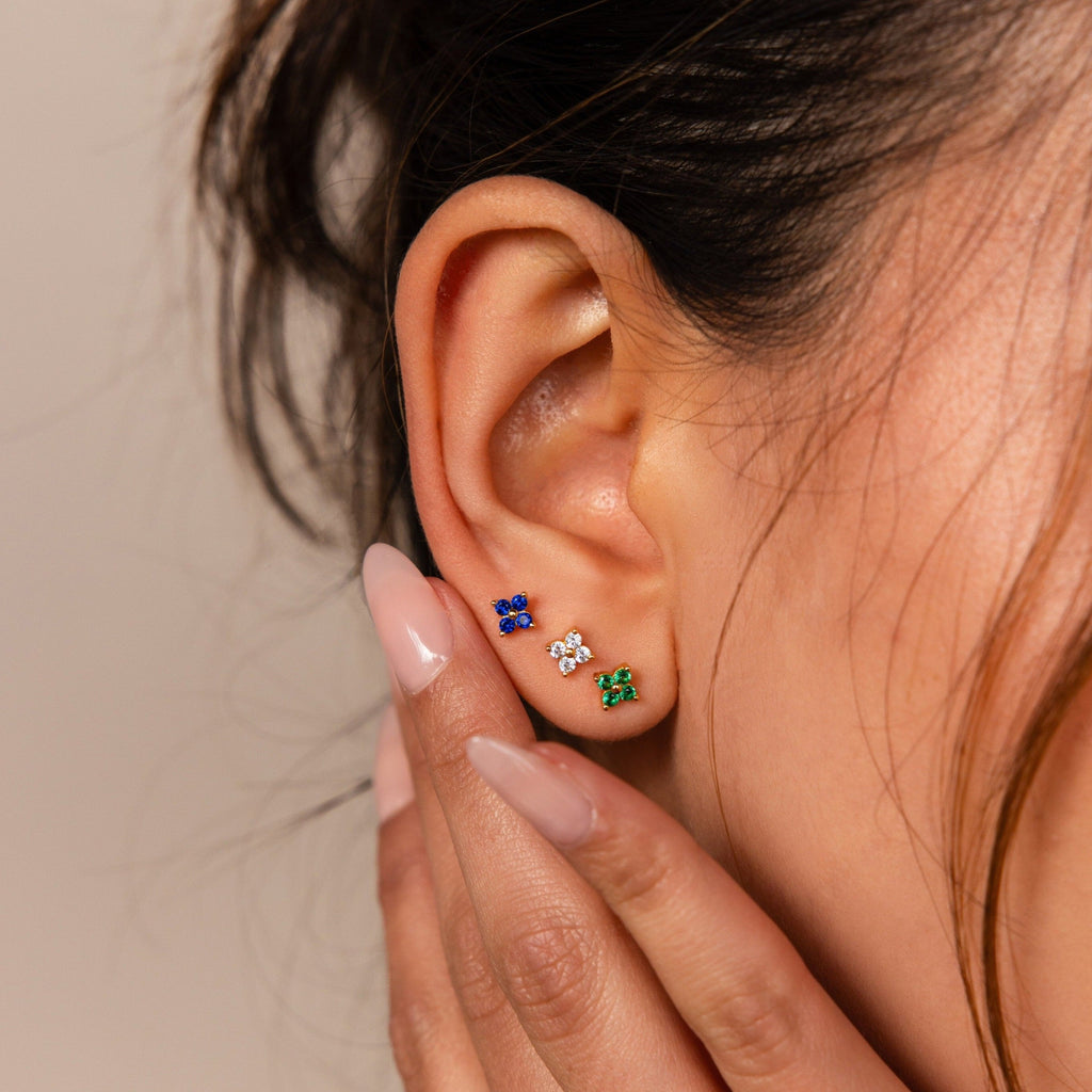 A close-up of a woman’s ear wearing the Flower Flat Back Studs Set, featuring three colorful gemstone stud earrings, including floral designs, with manicured nails touching her ear.