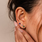 A close-up of a woman’s ear wearing the Flower Flat Back Studs Set, featuring three colorful gemstone stud earrings, including floral designs, with manicured nails touching her ear.