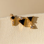 The Black Art Deco Earrings Set features three gold geometric earrings with black triangular stones and clear crystals, showcased on a textured cream surface—an elegant nod to minimalist jewelry.