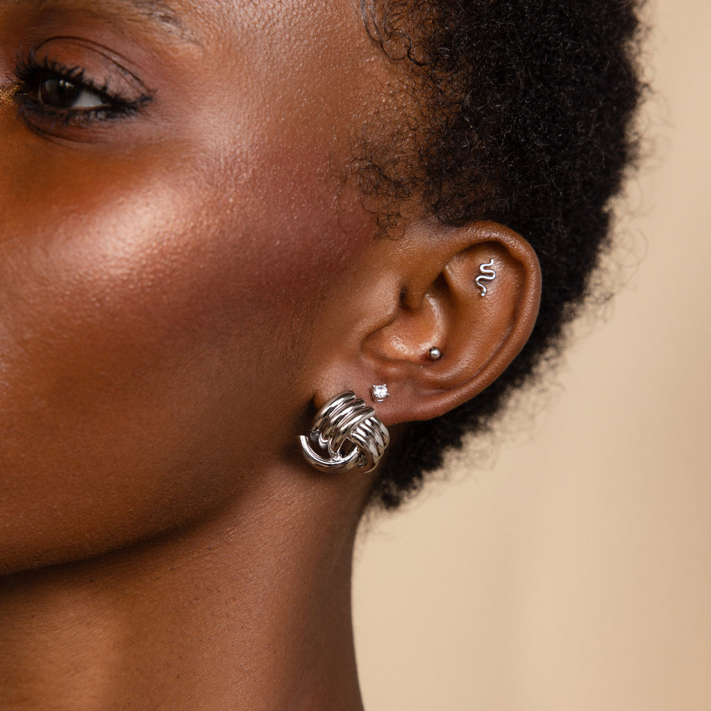Close-up of a person's ear adorned with multiple silver earrings, including a hoop, a wave-shaped earring, and the Bold Triple Knot Studs featuring a rope-like textured design for added dimension.
