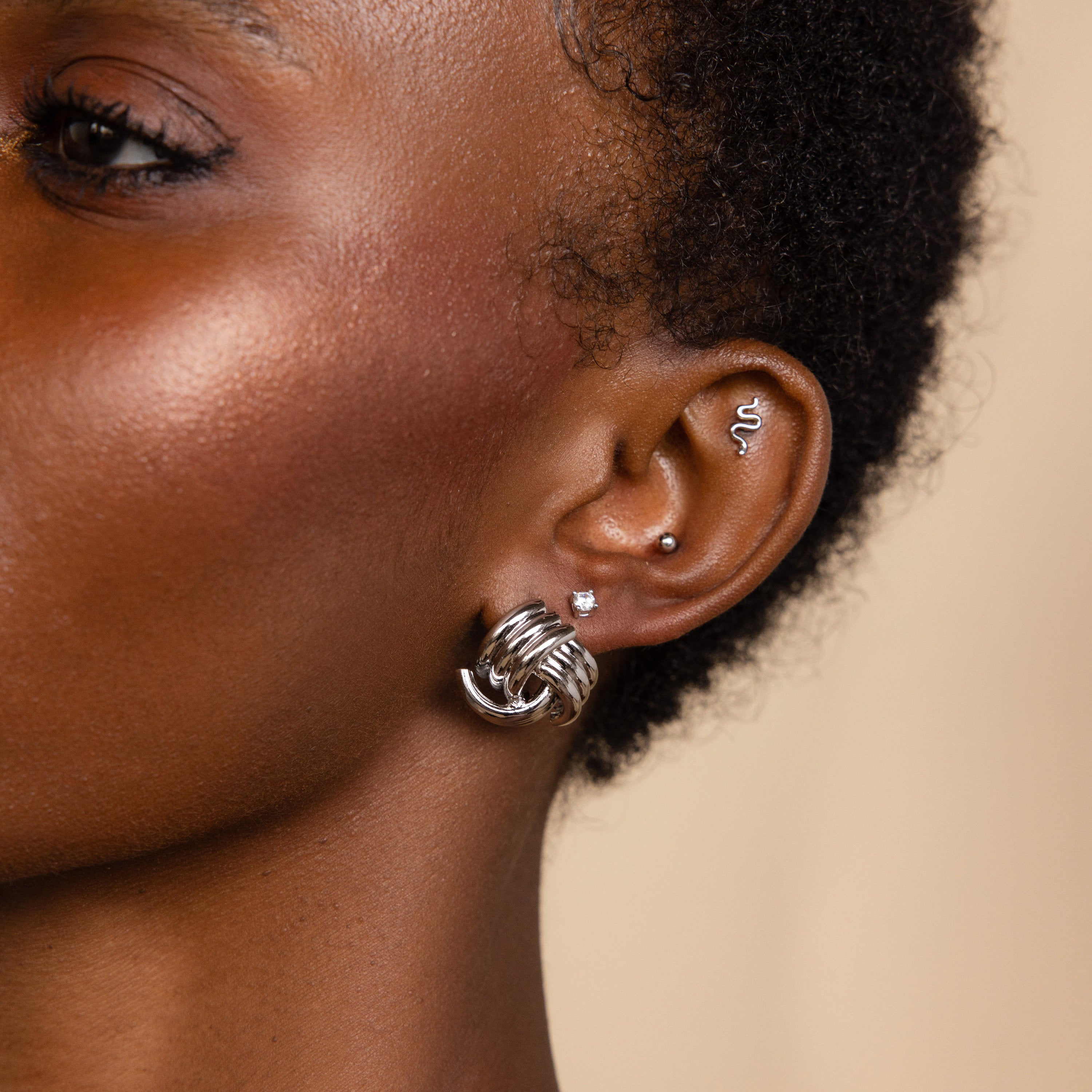 Close-up of a person's ear adorned with multiple silver earrings, including a hoop, a wave-shaped earring, and the Bold Triple Knot Studs featuring a rope-like textured design for added dimension.