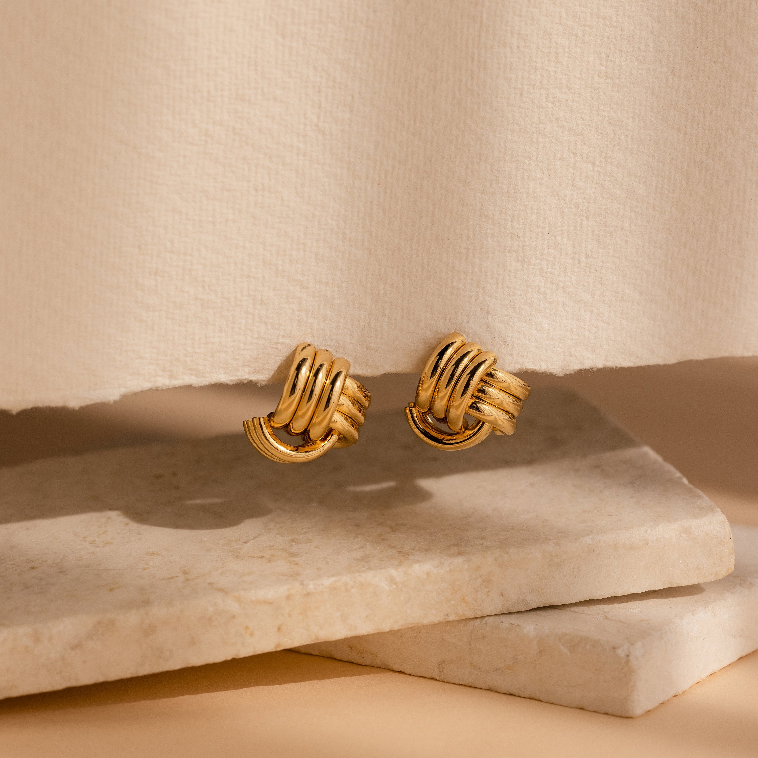 Bold Triple Knot Studs are showcased on textured stone slabs, highlighting their rope-like design against a neutral background.