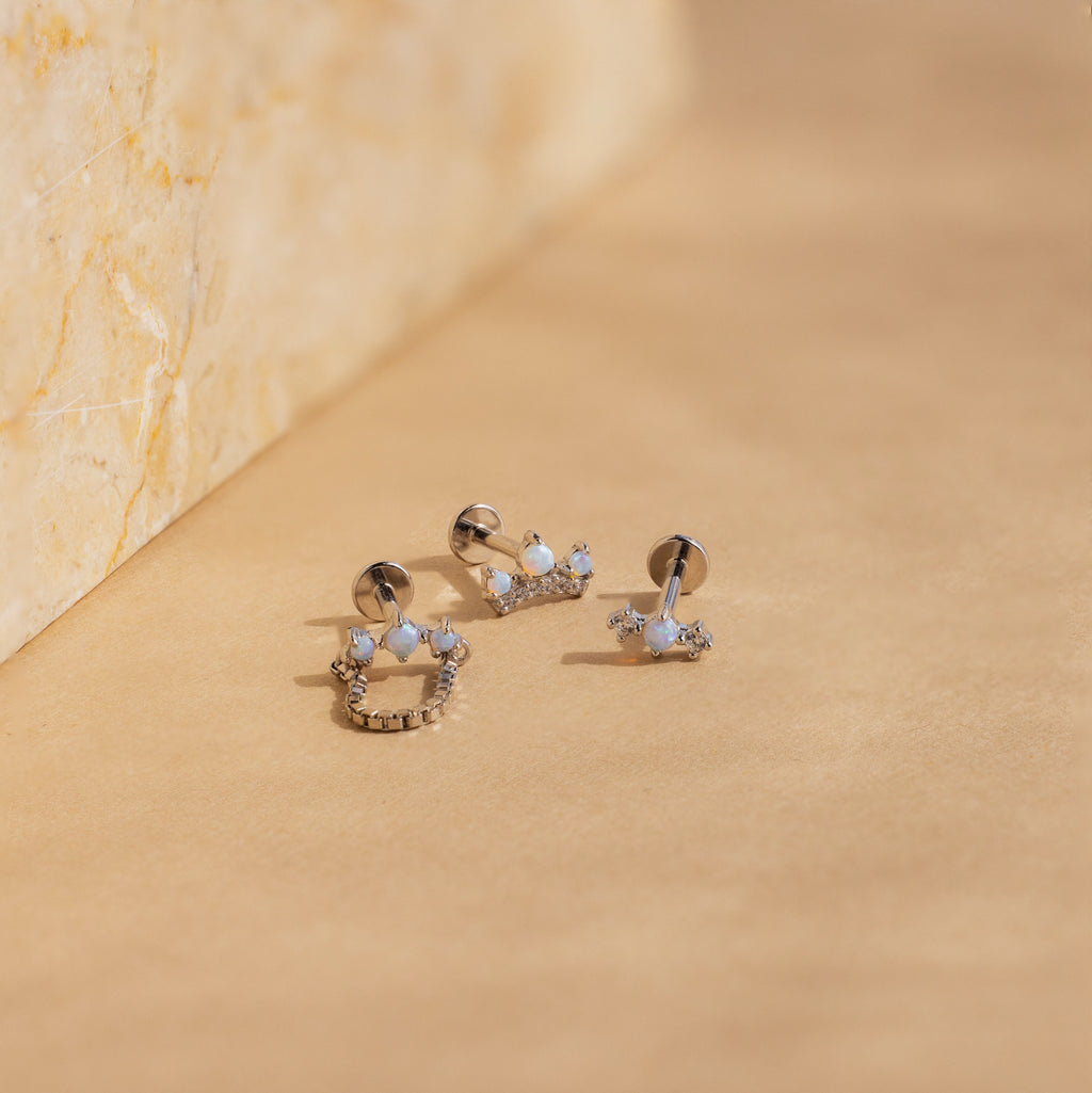 The Opal Cartilage Studs Set features three small silver earrings with opal, white, and clear stones displayed on a beige surface next to a marble block—ideal accents for any helix piercing.