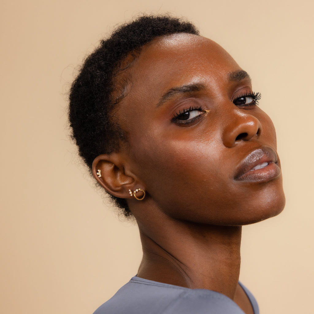 A woman with short hair looks confidently at the camera, wearing Opal Cartilage Studs Set earrings and a light blue top against a beige background.