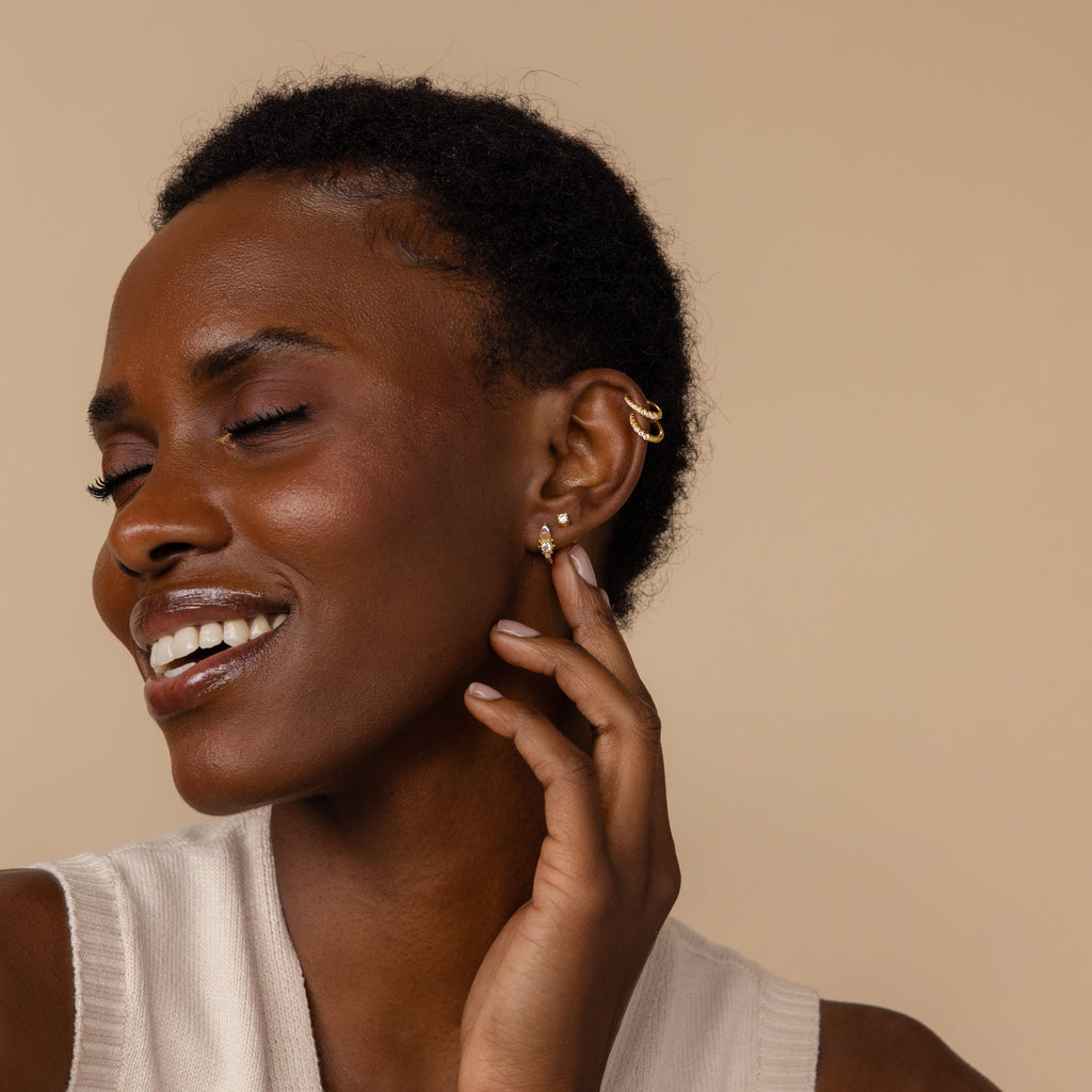 A smiling woman with short hair, in a cream sleeveless top, touches her face while wearing Opal and Diamond Huggies.