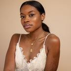 A woman wears a white lace top and gold layered necklaces, featuring the Sun & Moon Tarot Necklace, as she poses against a beige background.